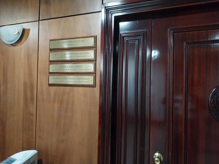 Rotulo en lat&oacute;n grabado en bajo relieve y patina de tinta negra, sobre metopa o peana de madera. Facultad de Medicina de la Universidad de Sevilla.