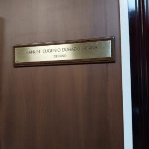 Rotulo en lat&oacute;n grabado en bajo relieve y patina de tinta negra, sobre metopa o peana de madera. Facultad de Medicina de la Universidad de Sevilla.