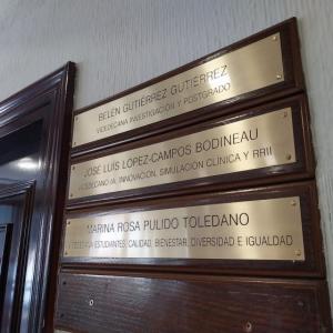 Rotulo en lat&oacute;n grabado en bajo relieve y patina de tinta negra, sobre metopa o peana de madera. Facultad de Medicina de la Universidad de Sevilla.