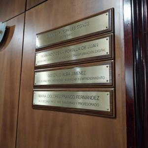 Rotulo en lat&oacute;n grabado en bajo relieve y patina de tinta negra, sobre metopa o peana de madera. Facultad de Medicina de la Universidad de Sevilla.